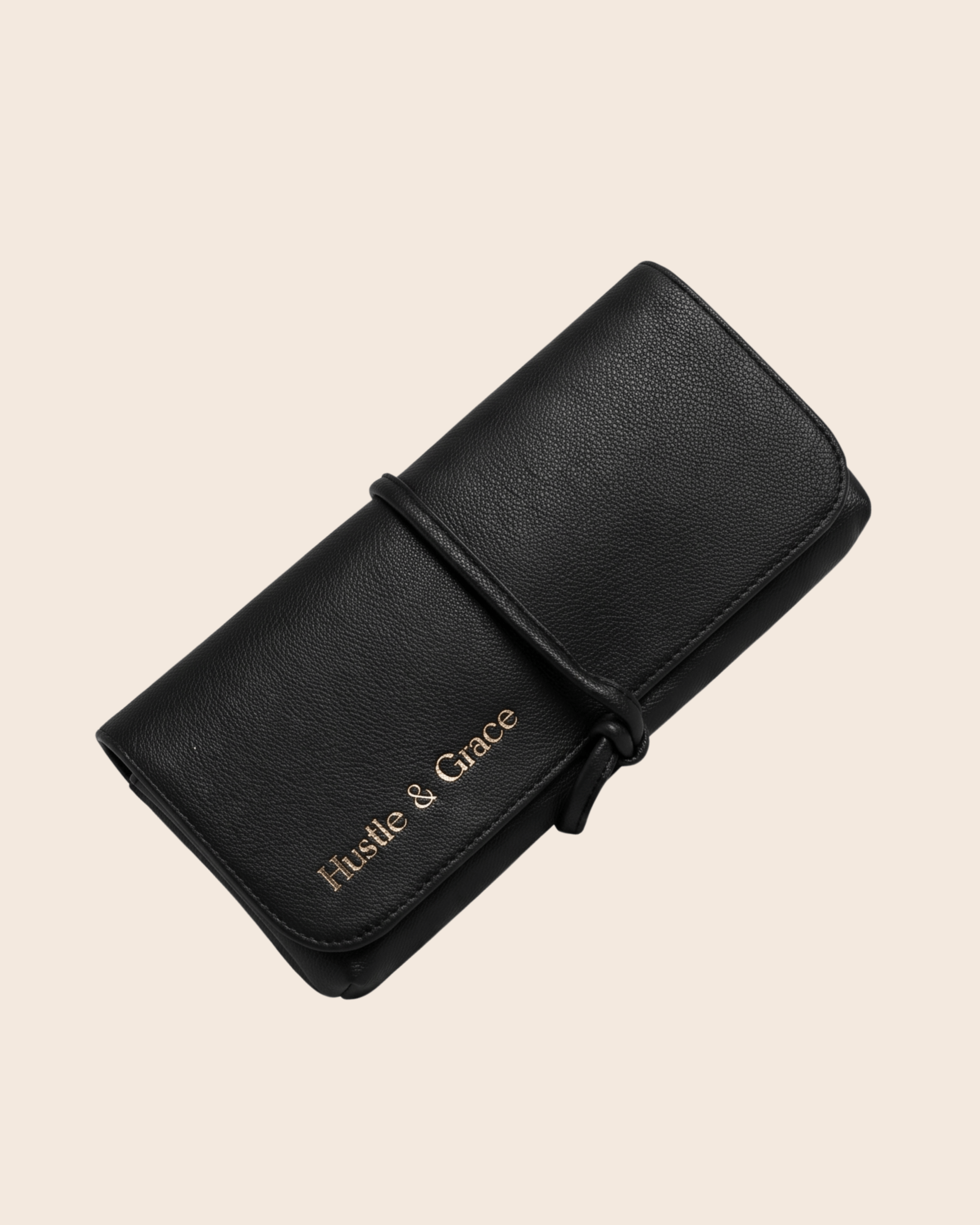A black pebbled vegan leather pouch or clutch with a wrap-around strap closure, featuring the brand name 'Hustle  & Grace' in gold lettering, shown on a solid light beige background.